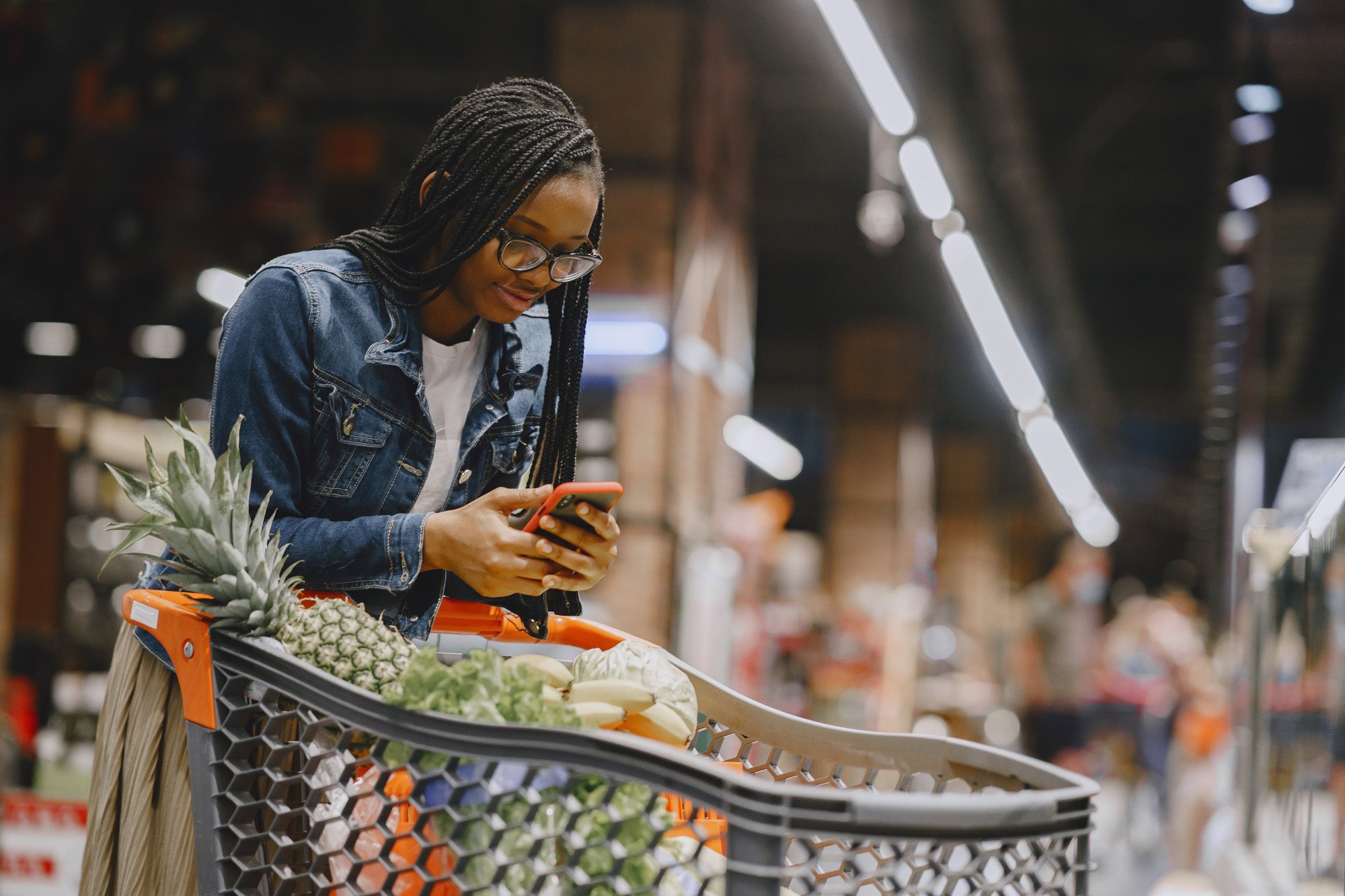 A shopper checking her phone beside a loaded supermarket trolley